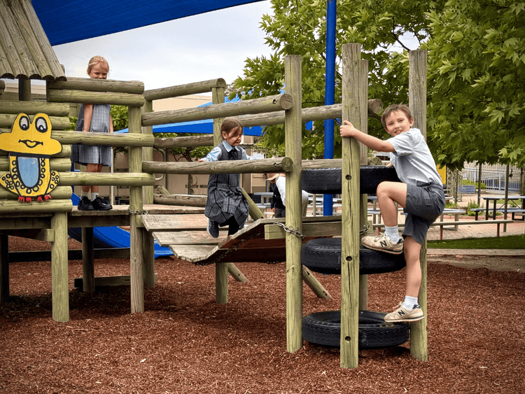 3 kids playing on school play equipment climbing on a tyre ladder, running across a wooden bridge