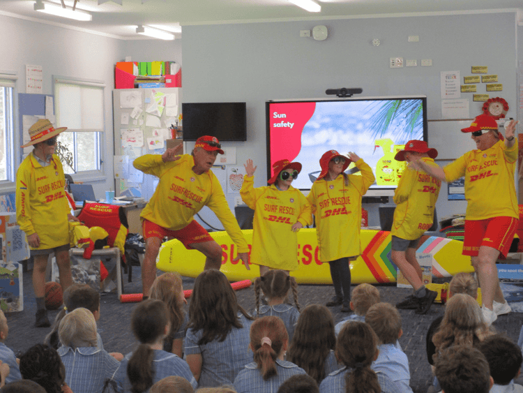 students watching a demonstration by two volunteer surf life savers. 3 students are dressed as surf life savers